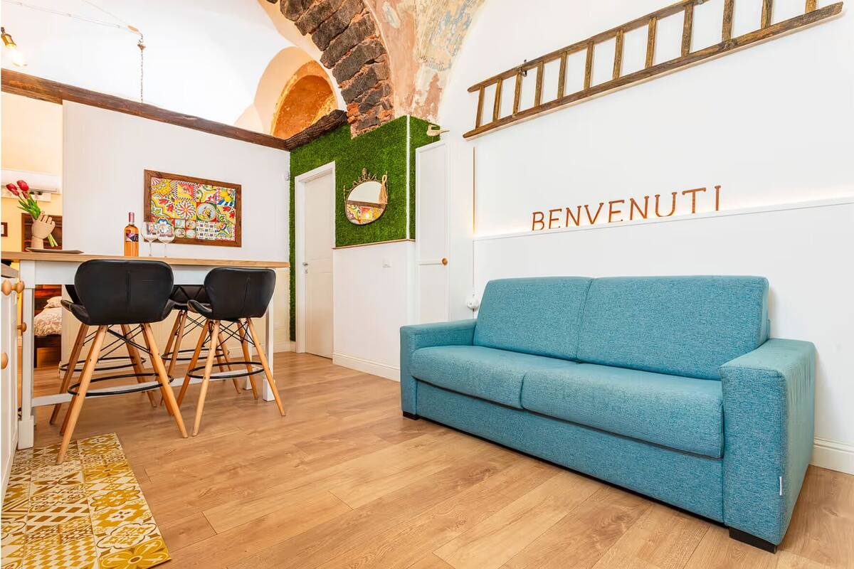 A modern living room with a blue sofa, wooden flooring, and a 'BENVENUTI' sign on the wall.