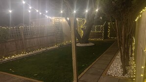A nighttime outdoor area with a paved walkway, a grassy lawn, and trees adorned with string lights. - Free Parking | Private Garden | Fully Equipped | Family Friendly | Bus Stop (Littlemore)