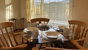 A dining area with a glass table, wooden chairs, and a view of a building through a window with sheer curtains. - Free Parking | Private Garden | Fully Equipped | Family Friendly | Bus Stop (Littlemore)