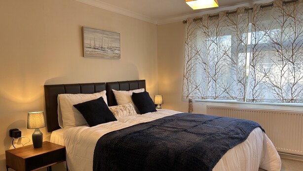 A neatly arranged bedroom with a bed, bedside tables, a lamp, a window with a view, and a painting on the wall. - Free Parking | Private Garden | Fully Equipped | Family Friendly | Bus Stop (Littlemore)