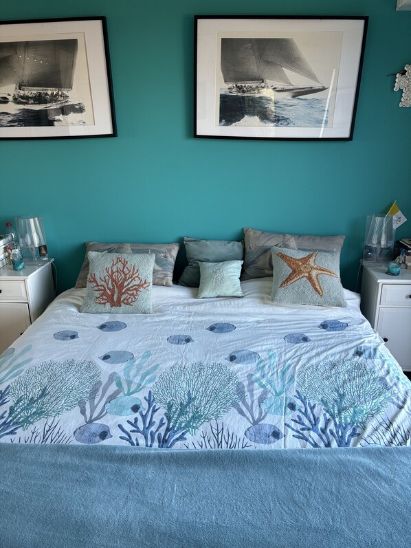 A bedroom with a bed featuring a blue and white ocean-themed bedspread, multiple pillows, and framed nautical artwork on the wall.