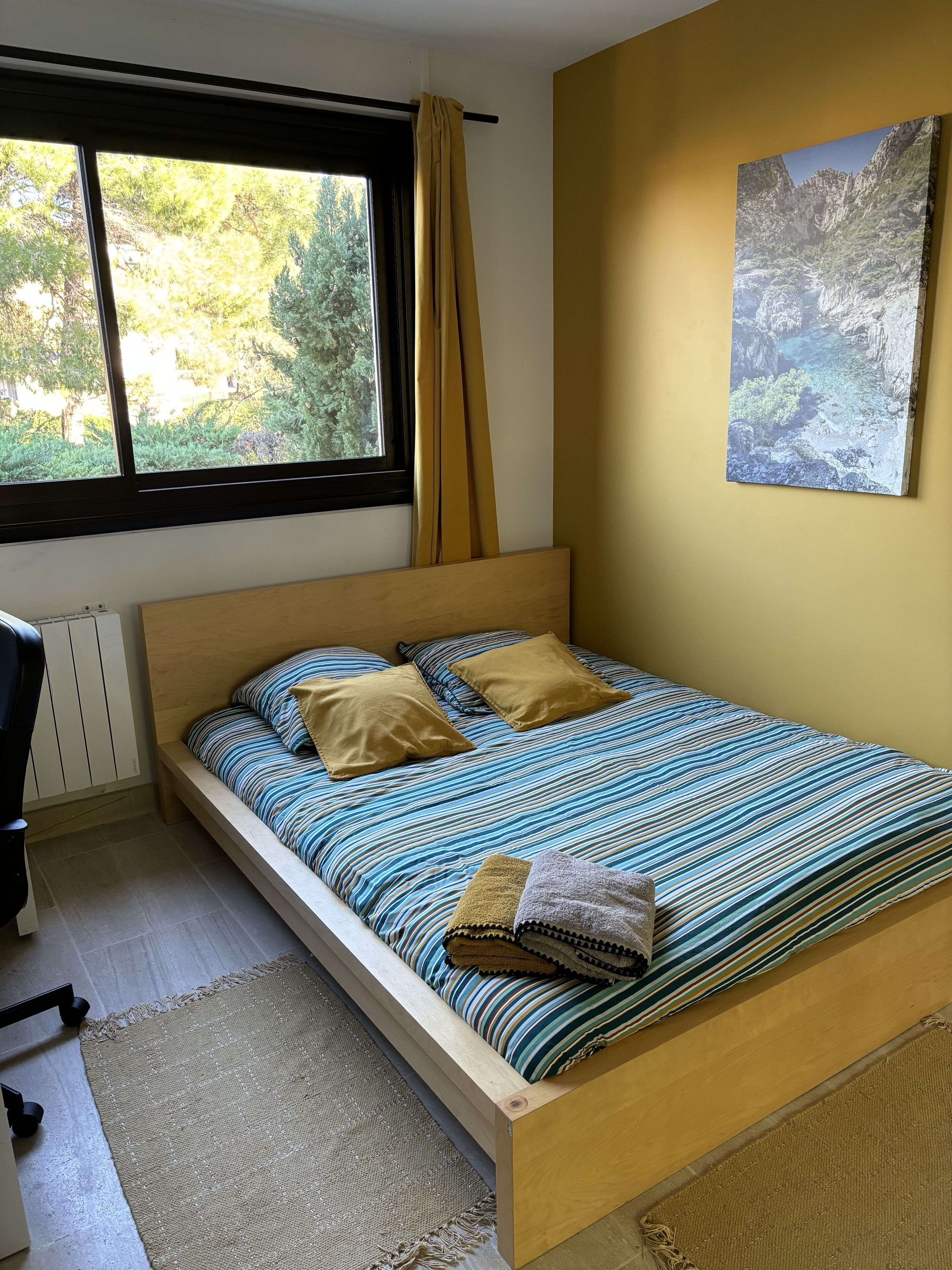 A bedroom with a bed, a window with curtains, a radiator, a wall-mounted painting, and a view of greenery outside.