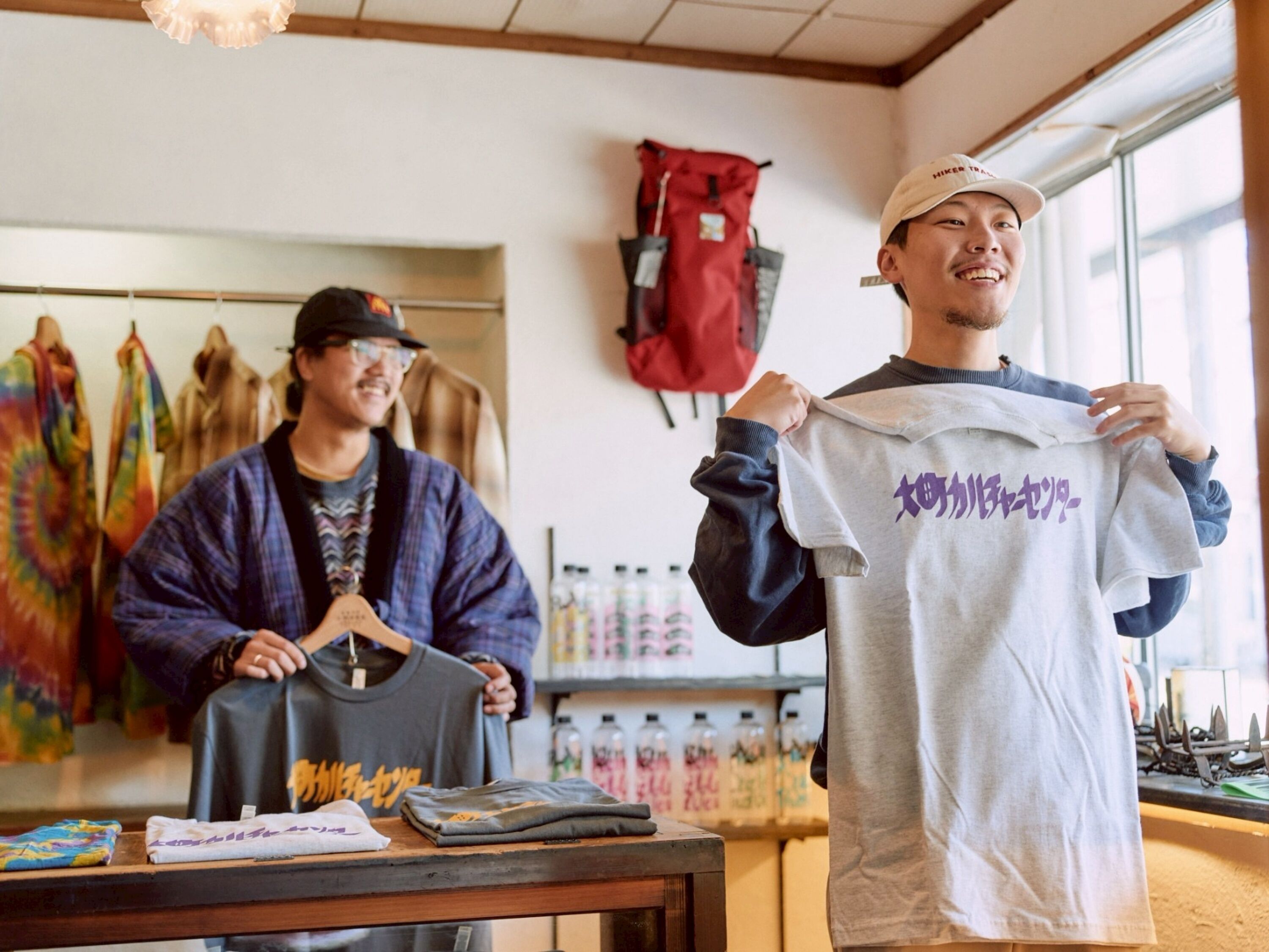 A person holding a grey t-shirt with Japanese text, another person holding a black bag, and a table with folded clothes and a colorful tie-dye shirt.