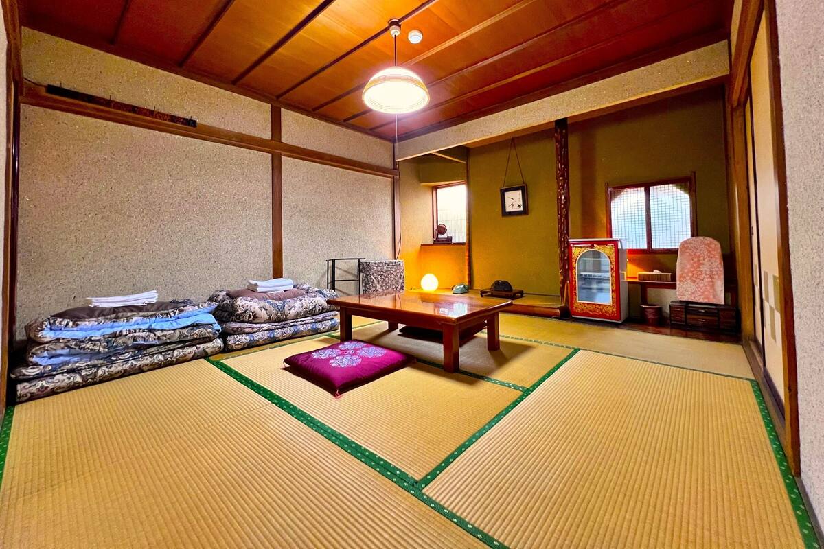 Ein traditioneller japanischer Raum mit Tatami-Böden, einem niedrigen Holztisch, einer Couch mit Kissen und einer verschiebbaren Tür zu einem weiteren Raum.