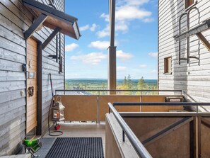 Un balcón de madera con barandilla metálica, vista a un paisaje boscoso y una escalera de metal que lleva a un nivel superior.