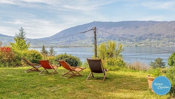 Een uitzicht op een meer met bergen op de achtergrond, twee ligstoelen en een bordje met de tekst 'Annecy Locatie'.