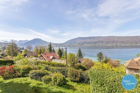 A scenic view of a lakeside property with houses, lush greenery, and mountains in the background.