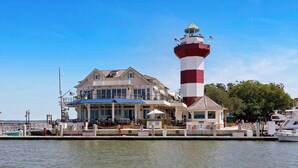 A waterfront building with a prominent lighthouse, a covered outdoor seating area, and a dock with boats. - Fairway One Villa Golf & Lagoon Views Walk to Beach Club (Hilton Head Island)
