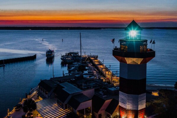 A lighthouse at dusk with a dock extending into the water, a boat, and a building with a sloped roof.