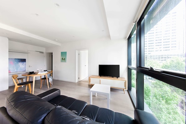 A modern living room with a black leather sofa, a white coffee table, and a TV on a wooden stand. There is a dining area with a wooden table and chairs, and a large window offering a view of the city.