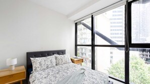 Ein Schlafzimmer mit großem Fenster, einem Bett mit floralem Bettbezug, einem Nachttisch mit Lampe und Blick auf Gebäude und Bäume.