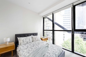 A bedroom with a large window, a bed with floral bedding, a bedside table with a lamp, and a view of buildings outside.