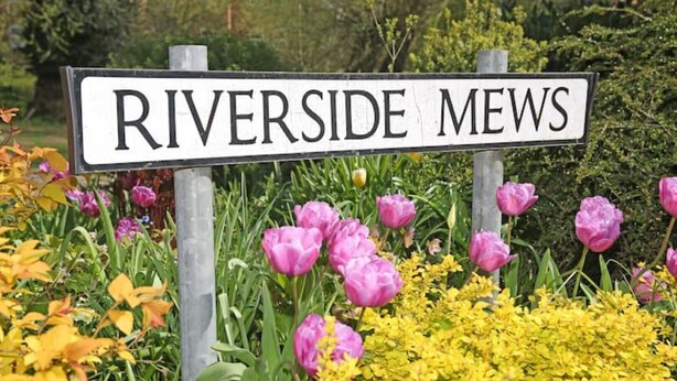 Un cartell que diu "RIVERSIDE Mews" envoltat de flors colorades.