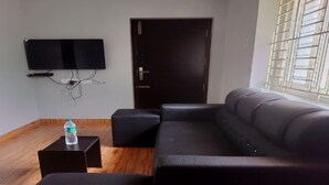 Una sala de estar con un sofá de cuero negro, un pequeño reposapiés negro, un televisor de pantalla plana montado en la pared y una ventana con persianas.