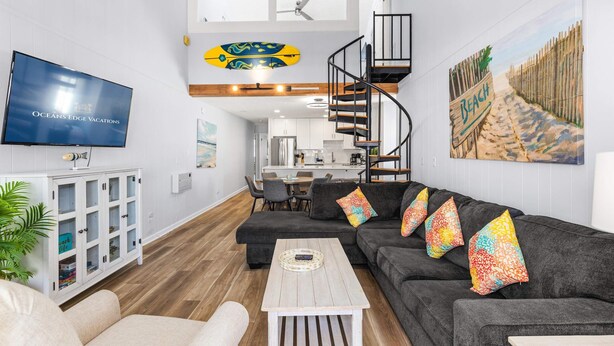 A modern living room with a large flat-screen TV, a dark grey sofa set, a white coffee table, and a spiral staircase.