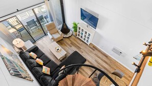 A modern living room with a black sofa, a wooden spiral staircase, and a TV mounted on the wall.