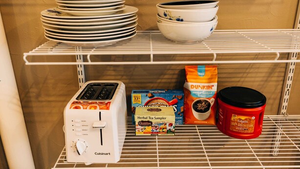 A kitchen shelf with stacked plates, bowls, a toaster, a box of herbal tea sampler, a bag of Dunkin' coffee, and a red mug.