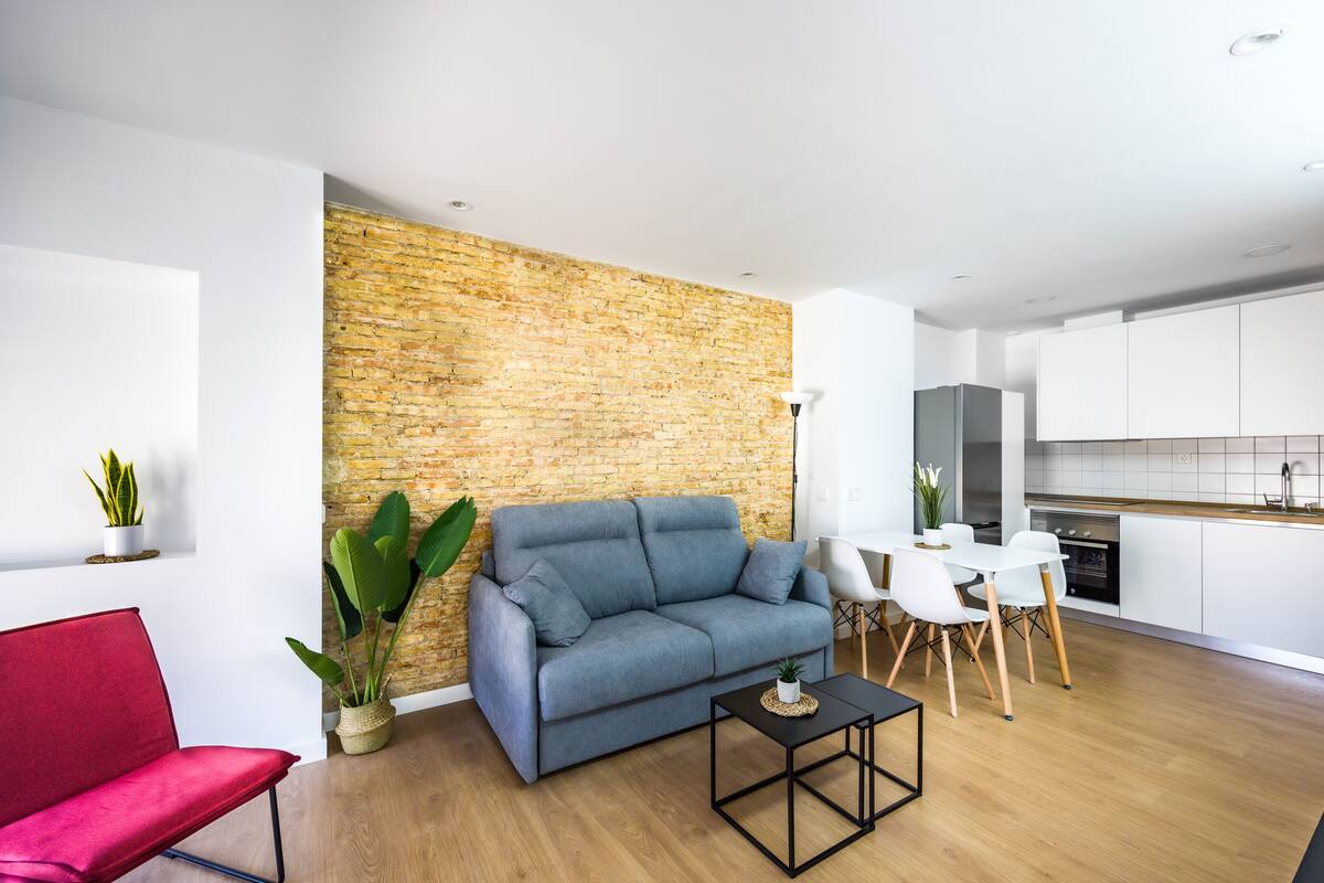 A modern living room with a blue sofa, a red chair, a small table, and a kitchen area with white cabinets and a brick wall.