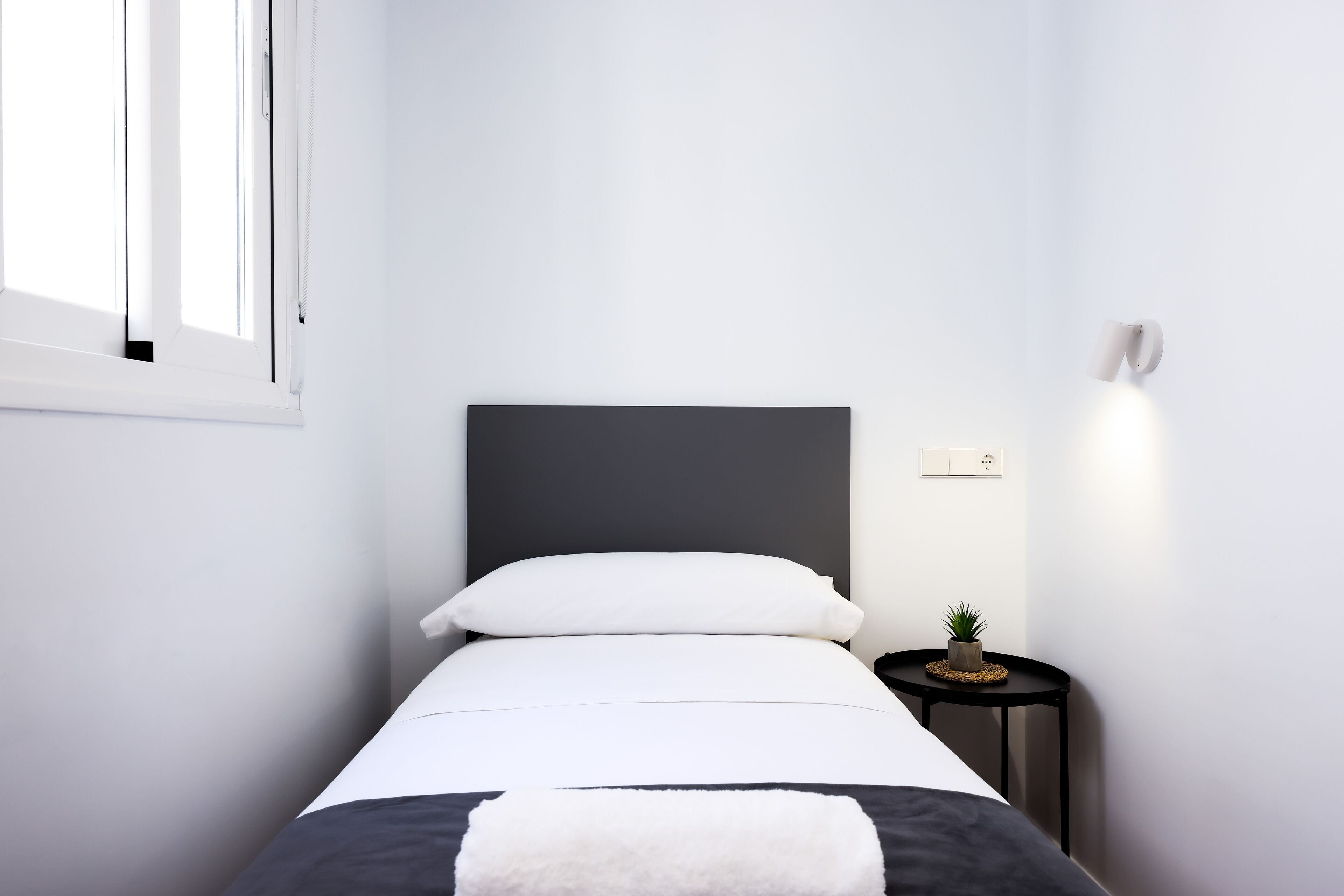 Un dormitorio minimalista con una cama, una mesita de noche con lámpara y una pequeña planta en maceta.