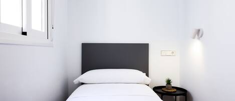 Un dormitorio minimalista con una cama, una mesita de noche con lámpara y una pequeña planta en maceta.