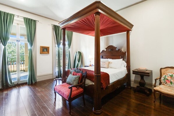 Un lit à baldaquin à quatre poteaux, doté d’une structure en bois et d’un baldaquin rouge, est placé dans une pièce au plancher en bois, où se trouvent également un fauteuil avec un coussin rouge et une petite table sur laquelle sont posés des livres.