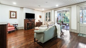 Ein geräumiges Wohnzimmer mit Holzdielen, Kamin, Fernseher und einem großen Fenster mit Blick ins Freie.
