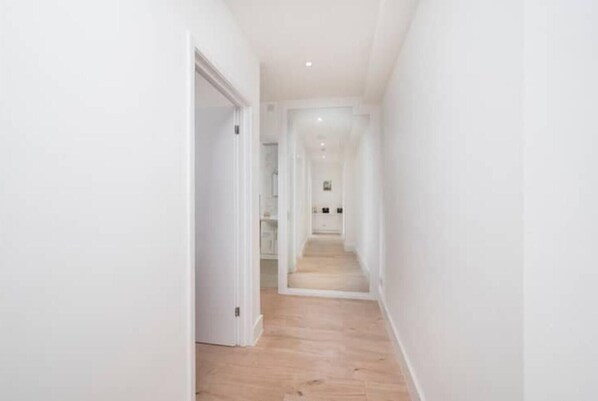 A narrow, light-colored hallway with wooden flooring leading to a room with white walls and a door.