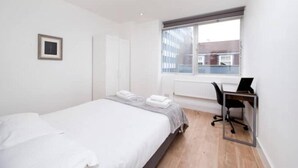 Una habitación de hotel moderna con una cama individual, un escritorio con una computadora portátil, una ventana con vistas a edificios y un radiador.