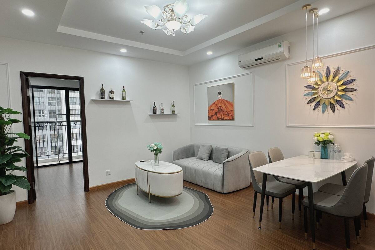 A modern living room with a grey sofa, a round coffee table, and a dining area with a white table and grey chairs. There is a large window with a view of buildings, and the room has a chandelier and wall-mounted air conditioning.
