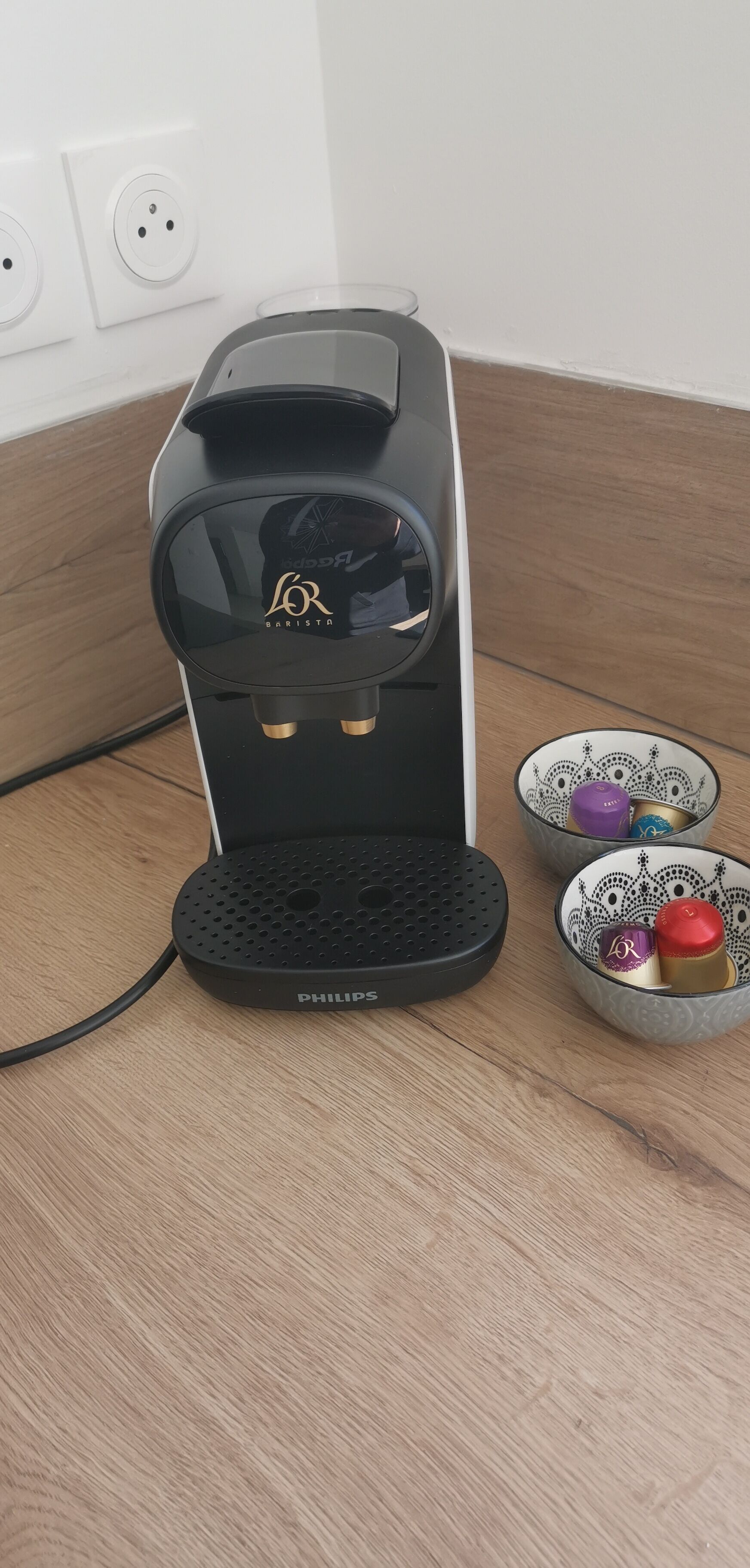 A black Philips coffee maker with a water reservoir and a control panel, placed on a wooden surface next to a decorative bowl with patterned lids.