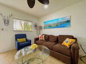 A living room with a brown sofa, a blue armchair, a glass coffee table, and a painting of a marina. - 3 miles to Fort Myers Beach with king bed (Fort Myers Beach)