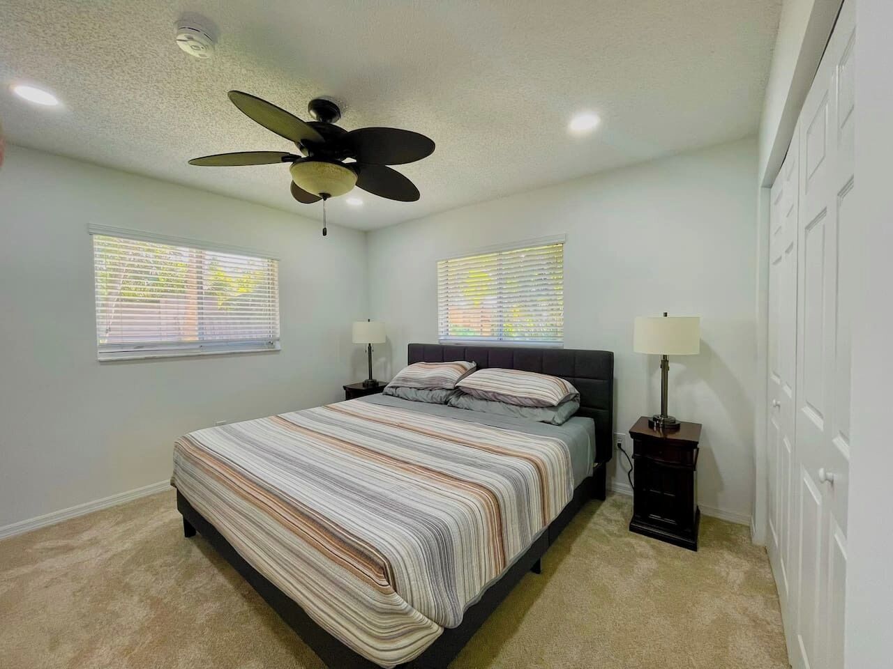 A bedroom with a bed, two bedside tables, a ceiling fan, and two windows with blinds.