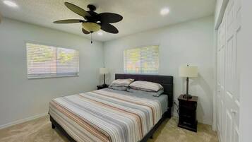 A bedroom with a bed, two bedside tables, a ceiling fan, and two windows with blinds.