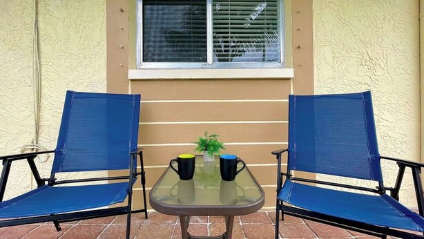 A patio with two blue chairs and a glass table, a small potted plant, and a window with blinds.