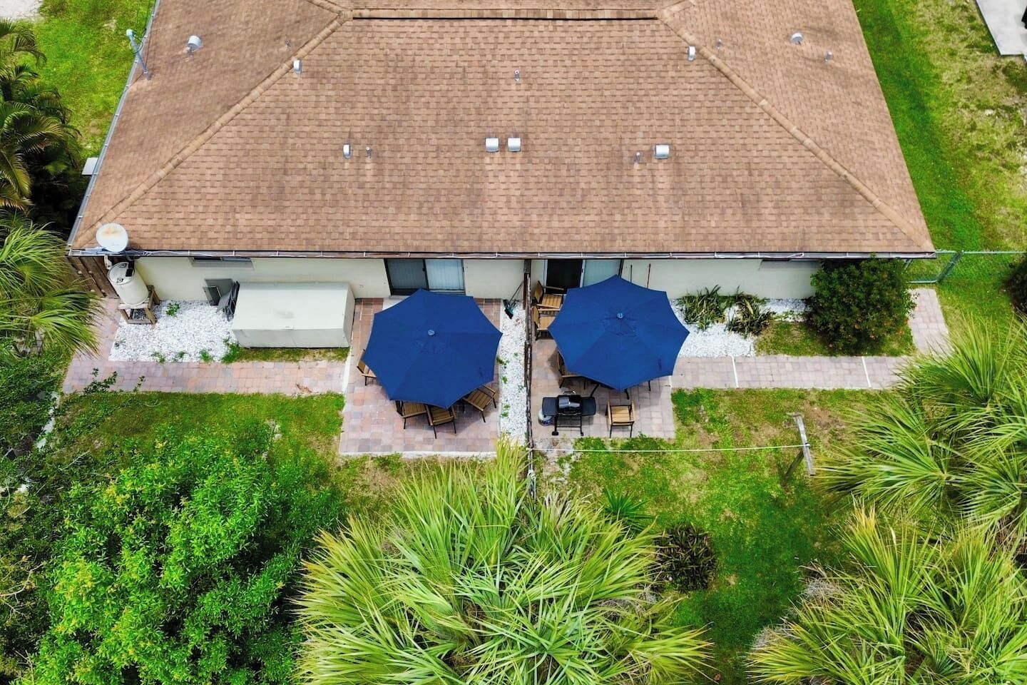 A two-story building with a tiled roof, a patio with two umbrellas, and a garden area with various plants.