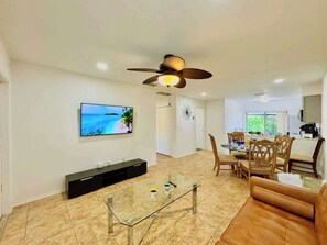 Ein modernes Wohnzimmer mit Fernseher, Glastisch, braunem Ledersofa, Essbereich mit Stühlen und Deckenventilator.