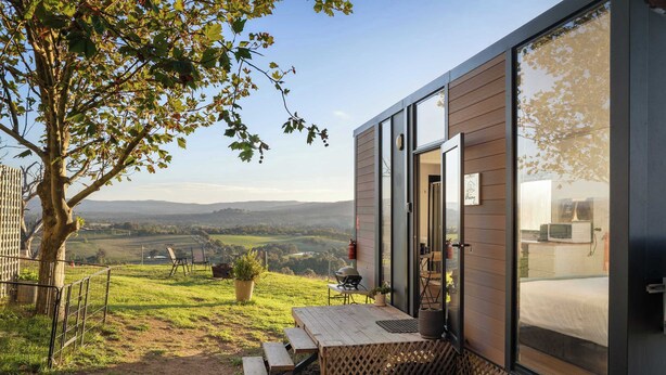 A modern cabin with a wooden exterior, large windows, a patio with a table and chairs, and a view of the countryside.