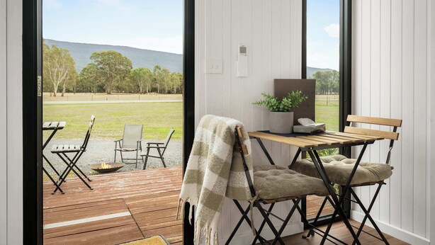 Una habitación con una terraza de madera, una mesa con sillas, un banco y vistas a un campo cubierto de césped y colinas.