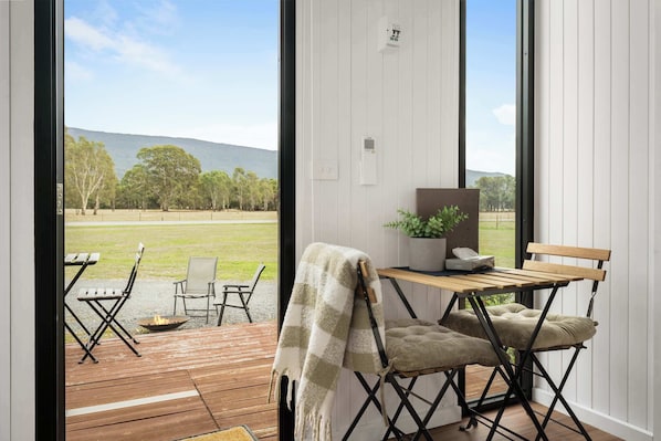 A room with a wooden deck, a table with chairs, a bench, and a view of a grassy field and hills.