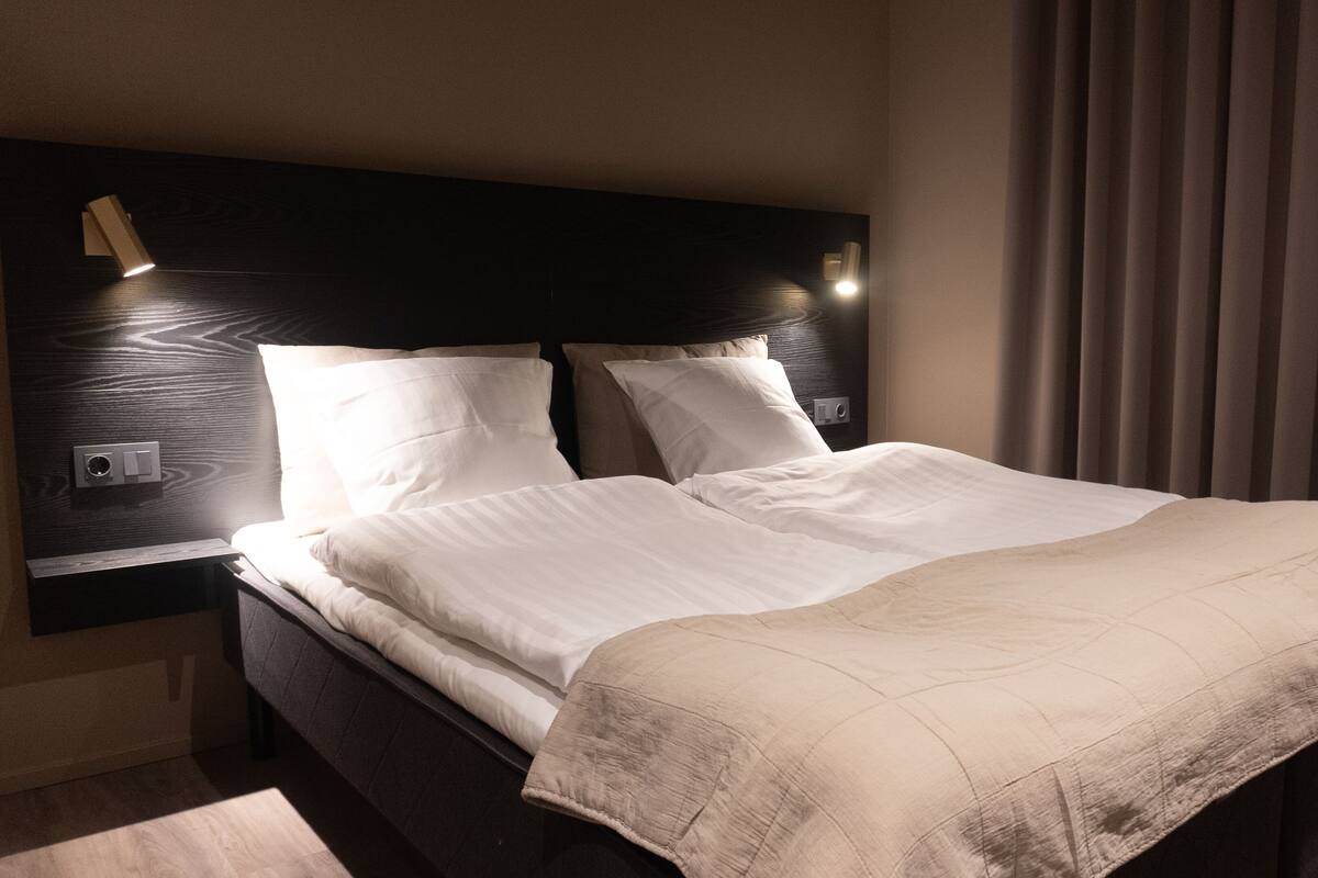 A neatly made bed with white linens and a beige throw blanket, a dark wooden headboard, and a wall-mounted light fixture.