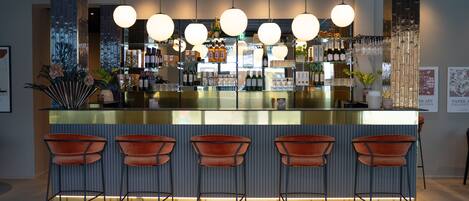 Un espace bar doté d’un comptoir en verre, de nombreuses bouteilles d’alcool et de plusieurs lampes suspendues rondes.