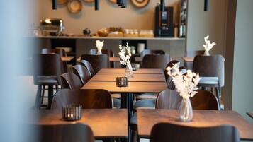 Interior de restaurante con varias mesas y sillas, un jarrón decorativo en el centro, y una barra con electrodomésticos de cocina al fondo.