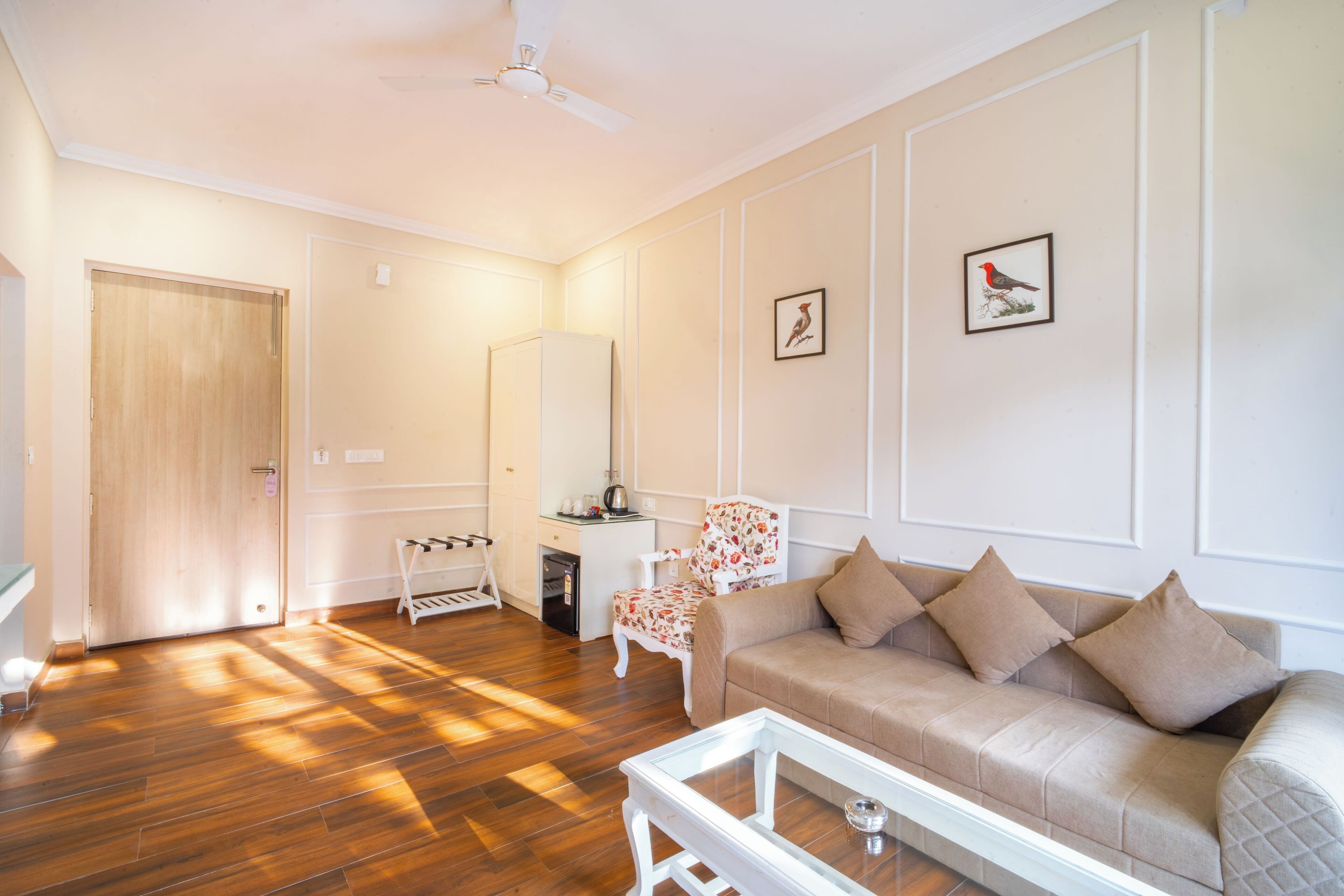 A living room with a wooden floor, a beige sofa, a white coffee table, a chair, a small table with a lamp, and a ceiling fan.
