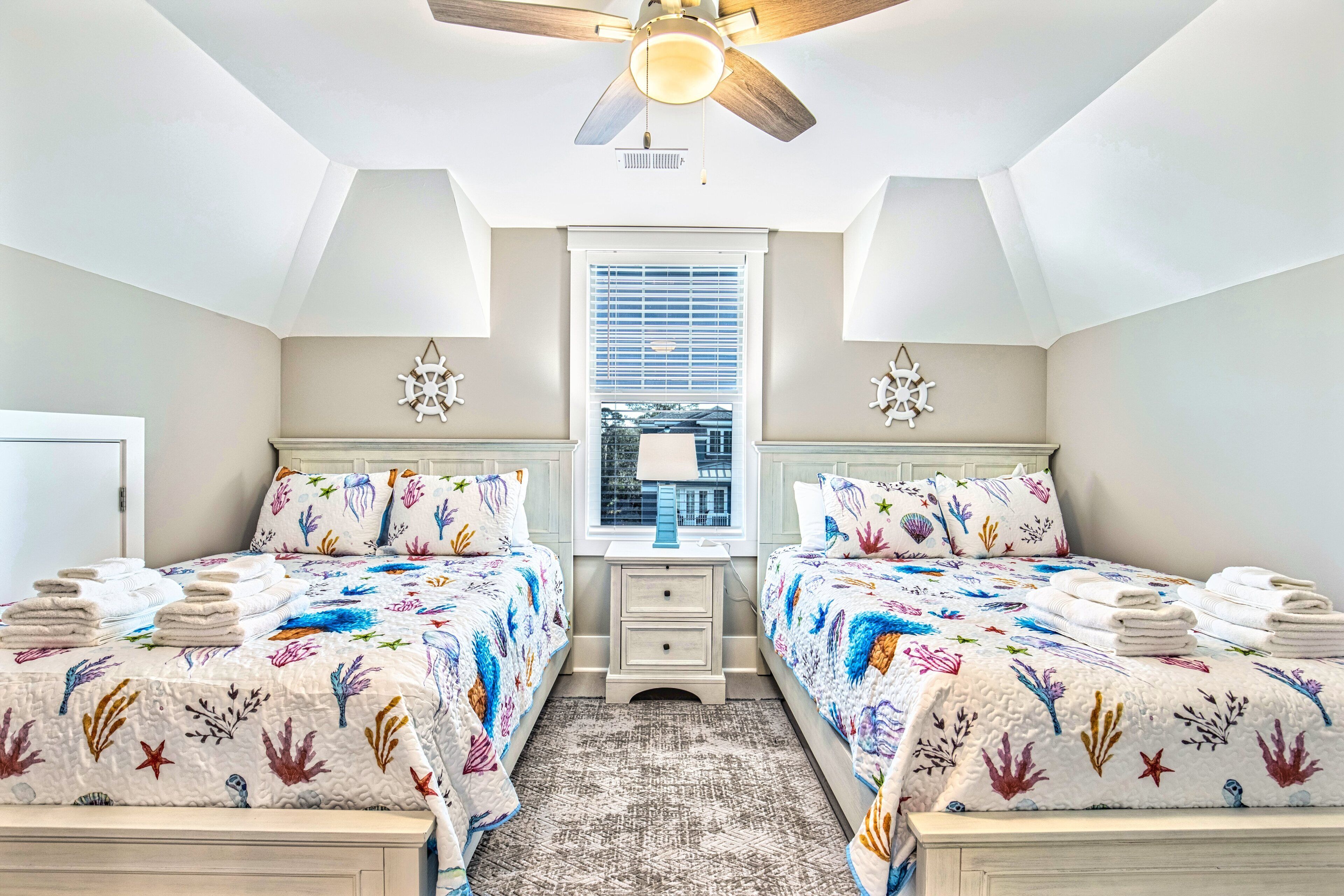 A bedroom with two beds, a ceiling fan, a window with blinds, and a view of a building outside.