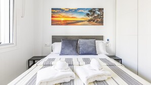 Una cama bien hecha con una colcha estampada y dos almohadas, un cuadro colgado en la pared que muestra un atardecer y una mesita de noche con una lámpara.