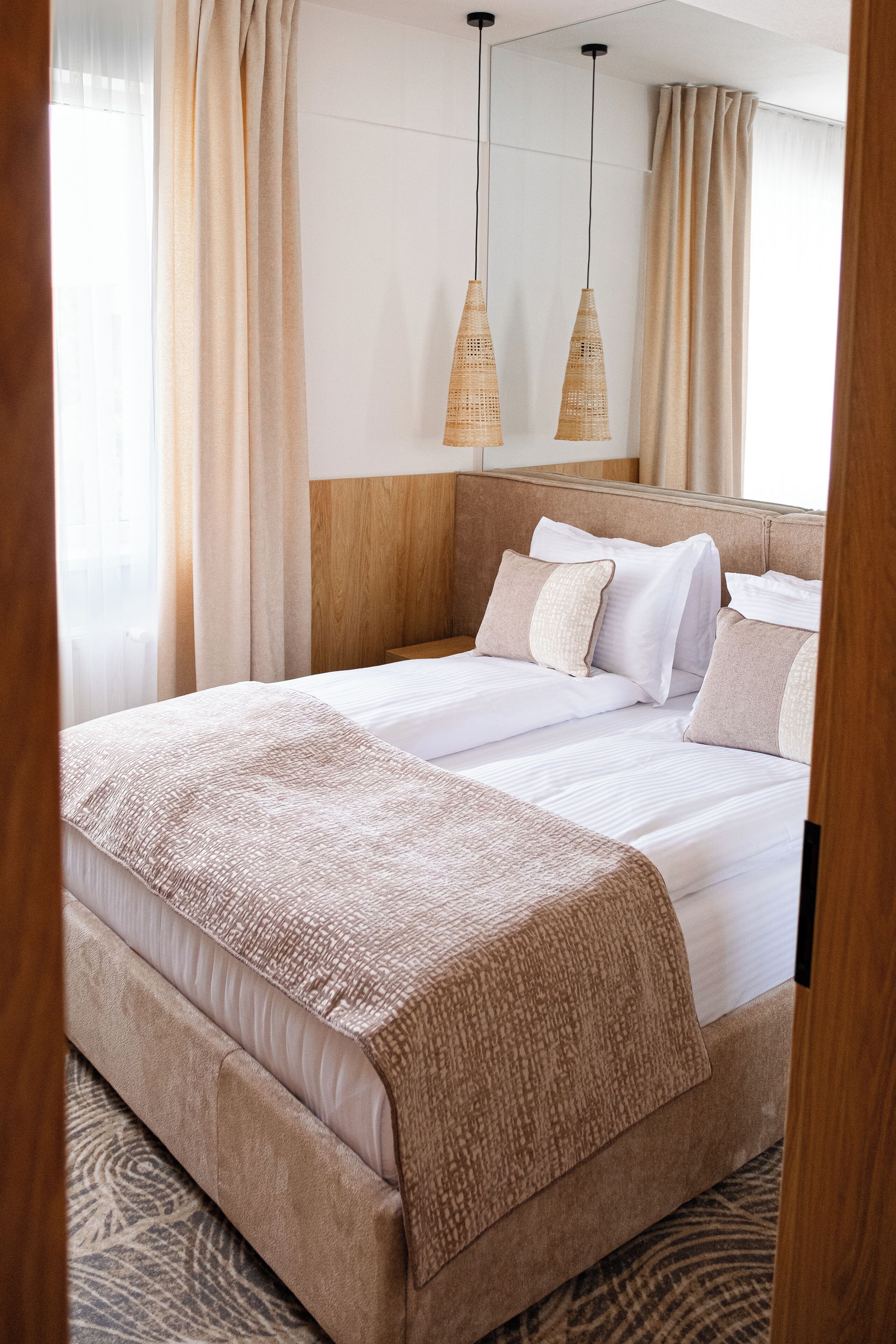 A neatly made bed with a beige headboard, white linens, and a textured throw blanket. There are two hanging wicker lamps above the bed, and a large mirror on the wall.