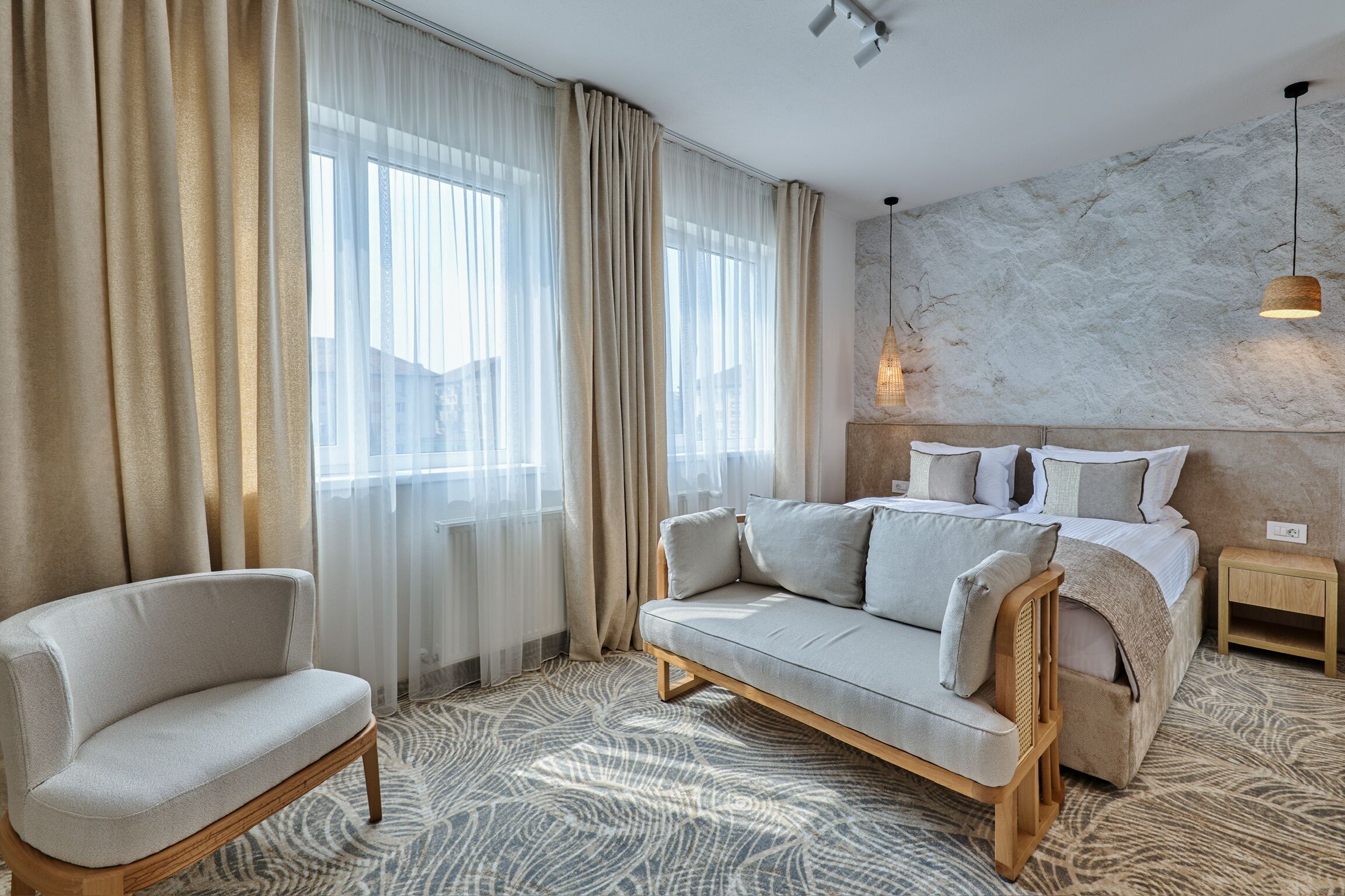 Una habitación de hotel moderna con sofá, una silla, una cama con cabecera de madera, una mesita de noche y un tapete estampado.