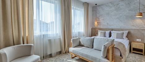 Una habitación de hotel moderna con sofá, una silla, una cama con cabecera de madera, una mesita de noche y un tapete estampado.