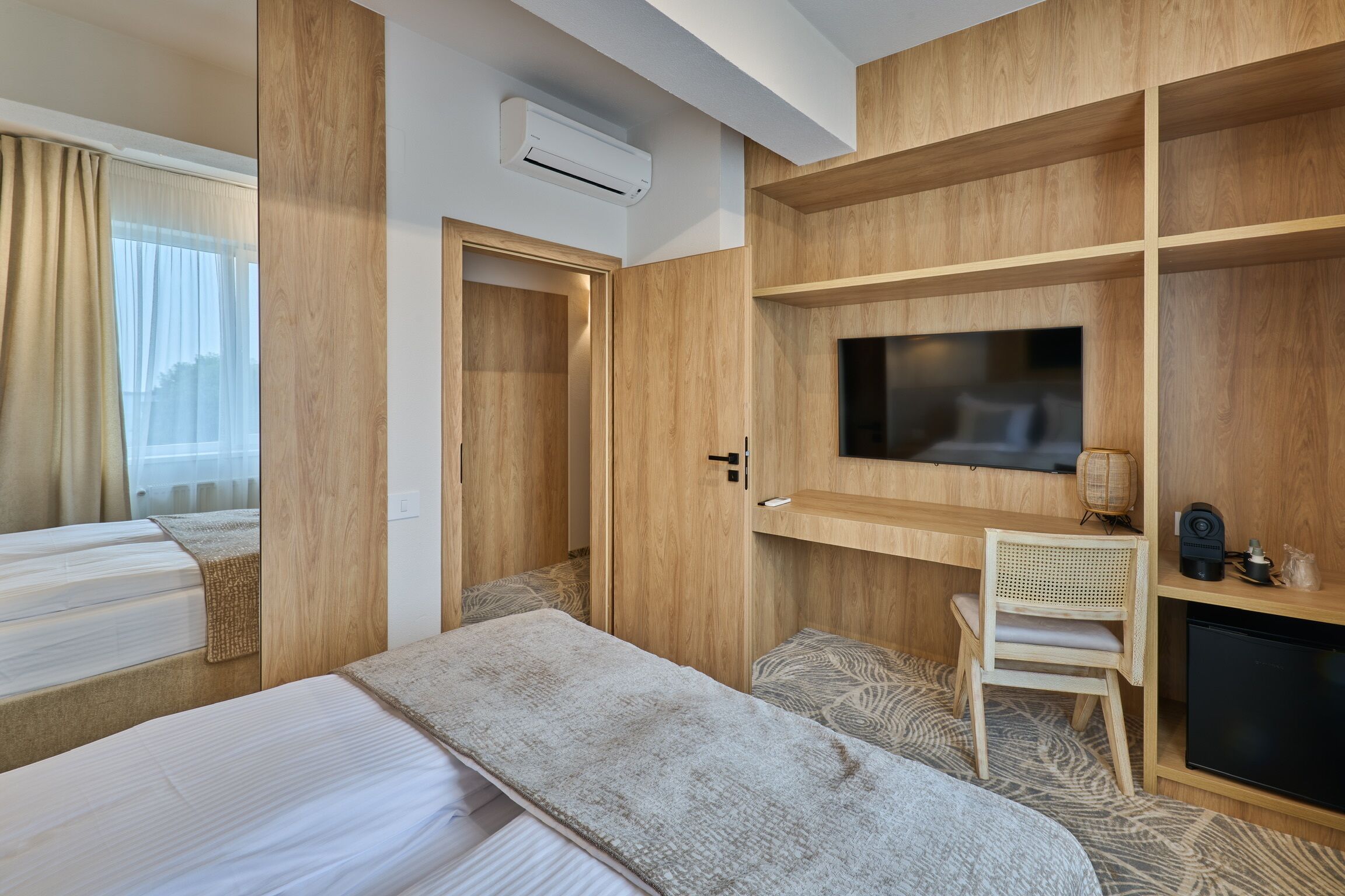 Habitación de hotel con cama, televisor montado en la pared, escritorio con silla y estanterías empotradas.