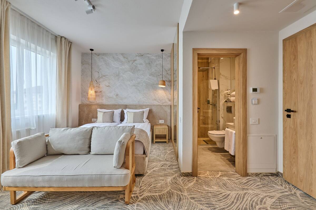 A modern hotel room with a sofa, a bed, a marble wall, a wooden door, and a bathroom visible through an open door.
